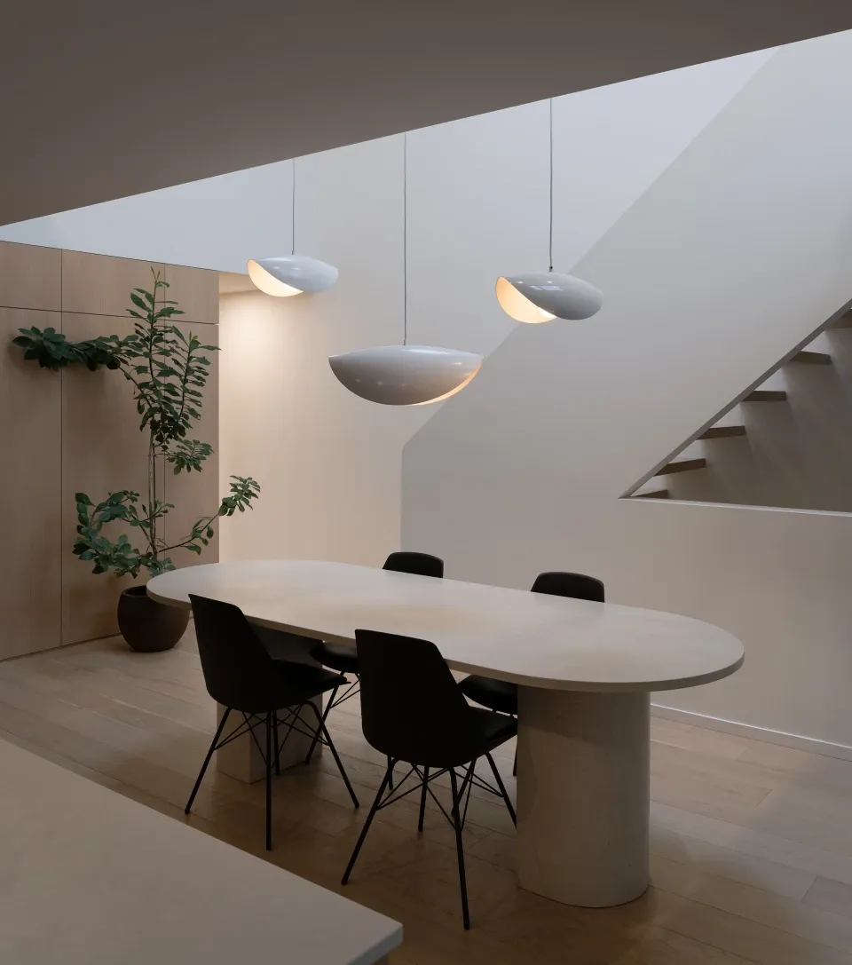 Luminaire Authentik Coquelicot pendant lights, a cluster of three staggered, curved white shell-like fixtures suspended over a modern white oval dining table with black chairs in a minimalist, light-filled space near a modern staircase.