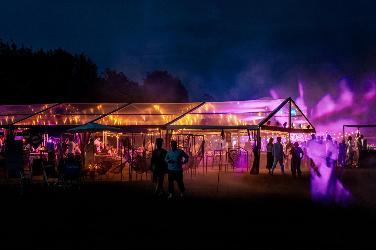 Outdoor evening event under a transparent tent with warm string lights and people socializing, illuminated by purple and orange lighting.