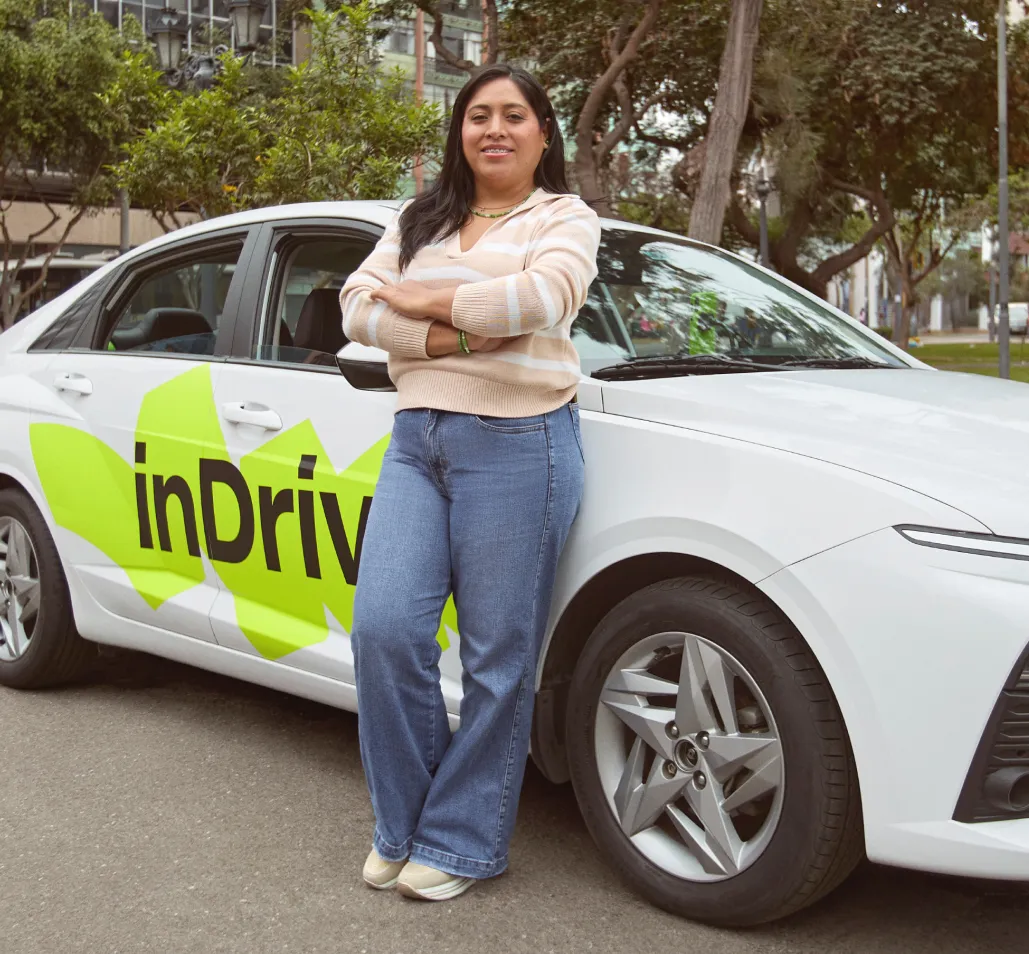 Woman in a beige striped sweater and blue jeans standing with arms crossed, leaning against a white car with bright green logo text 'inDrive' on its side, parked on a city street with trees and tall buildings in the background.