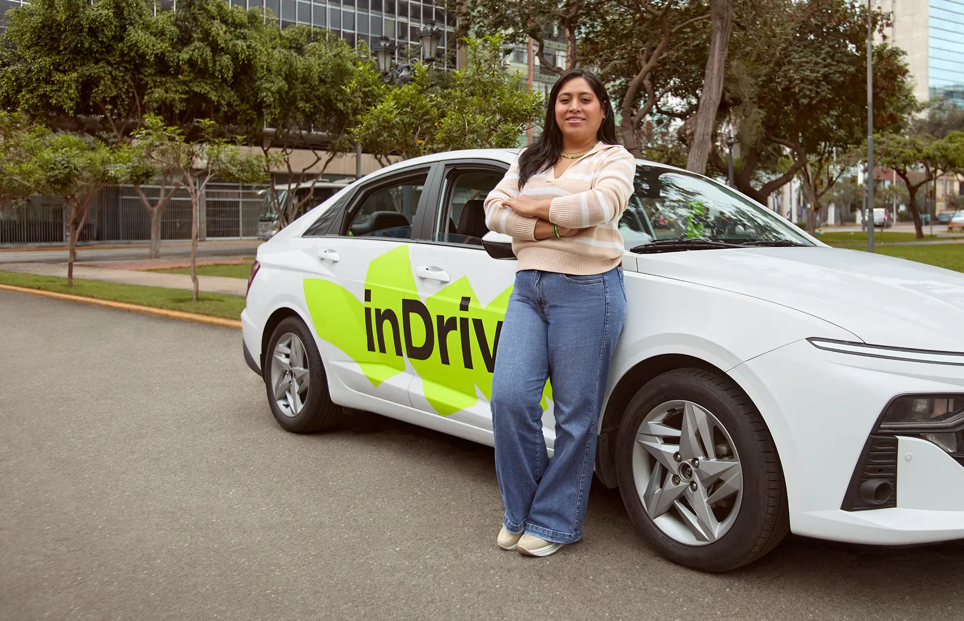 Woman in a beige striped sweater and blue jeans standing with arms crossed, leaning against a white car with bright green logo text 'inDrive' on its side, parked on a city street with trees and tall buildings in the background.