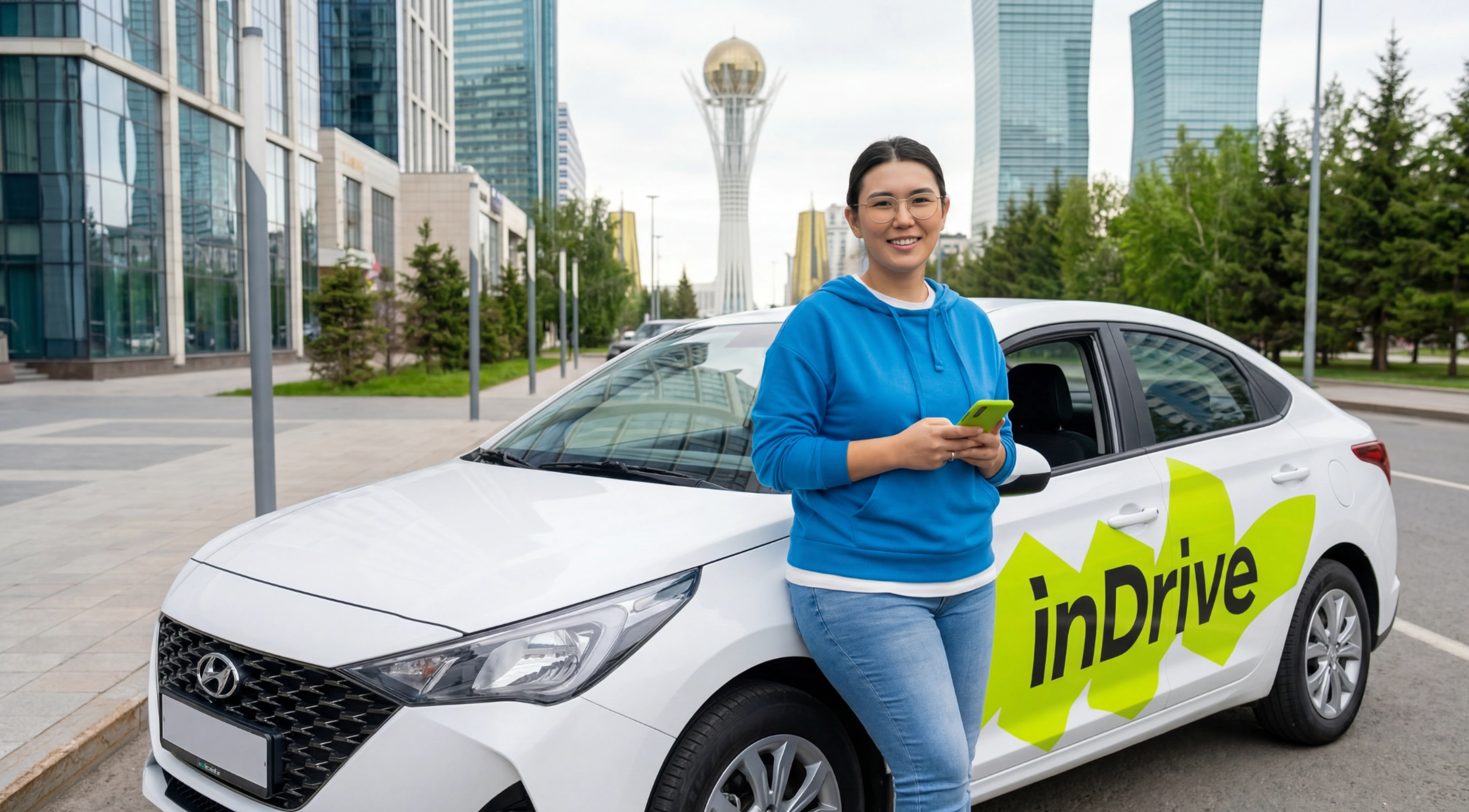 A smiling woman wearing glasses and a blue hoodie stands next to a white Hyundai car with a bright green 'inDrive' logo on the door, holding a green phone, with tall modern buildings and a distinctive tower in the background.
