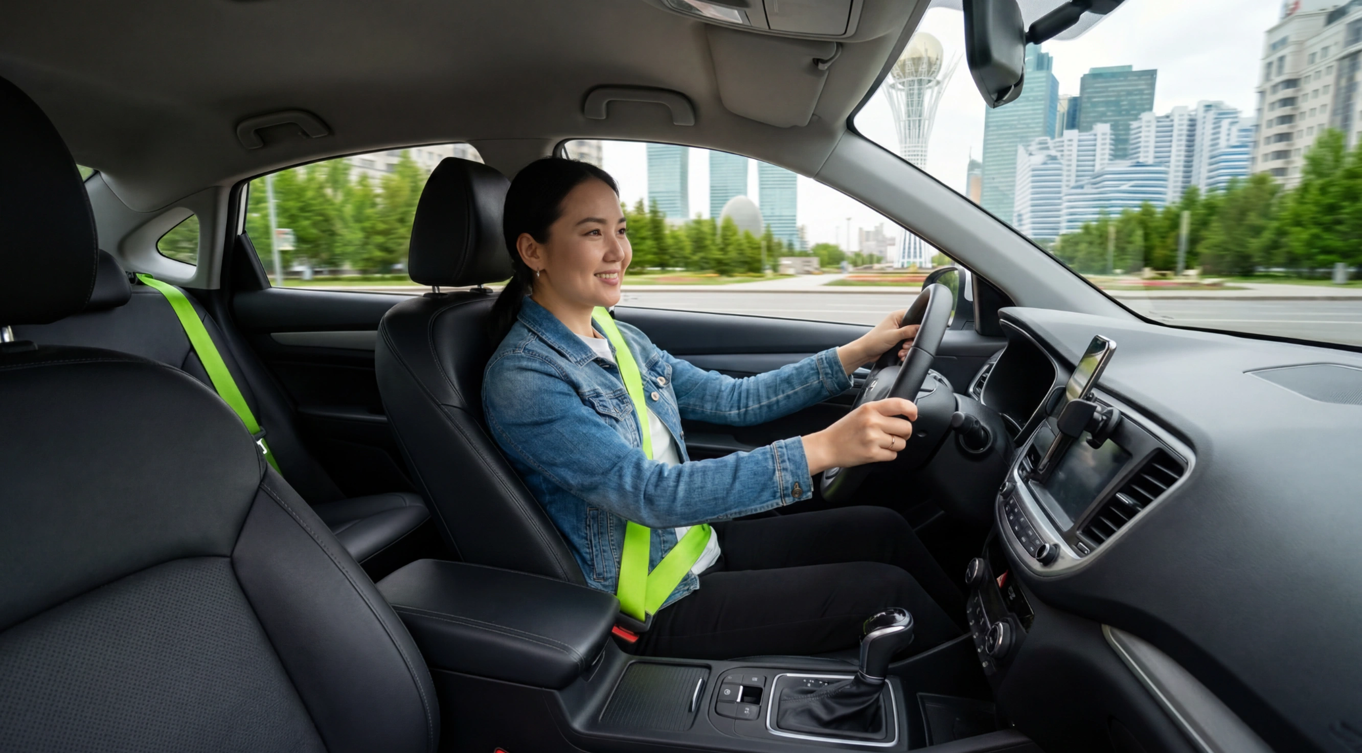 Smiling woman wearing a denim jacket and neon green seatbelt driving a modern car through a city with tall buildings and greenery visible outside.