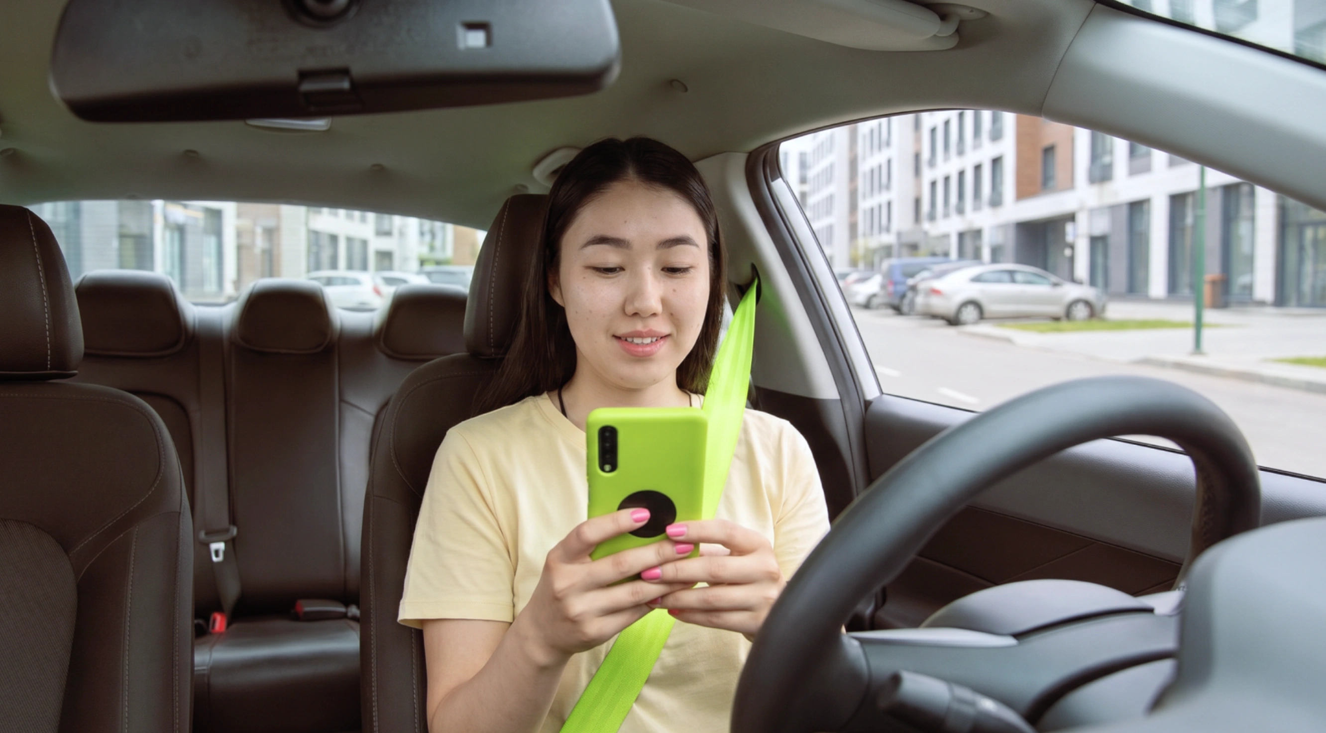 A young woman sitting in the driver's seat of a car, wearing a neon green seatbelt, looking down and smiling at her phone with a matching green case inside the car parked near modern buildings.