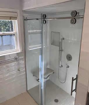 Modern glass shower enclosure with a wall-mounted folding seat, handheld showerhead, and large window beside it.