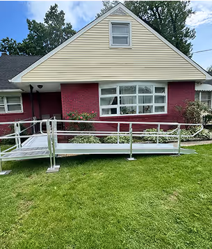 Metal wheelchair ramp installed in front of a single-story house with yellow siding and red brick lower facade.