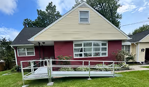 Single-story house with red brick lower walls and beige upper siding, featuring a metal wheelchair ramp leading to the front door.