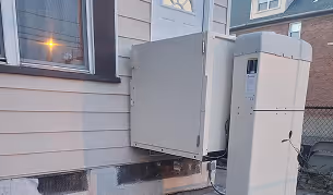 Outdoor HVAC units installed beside a building with light-colored siding and a nearby window at dusk.
