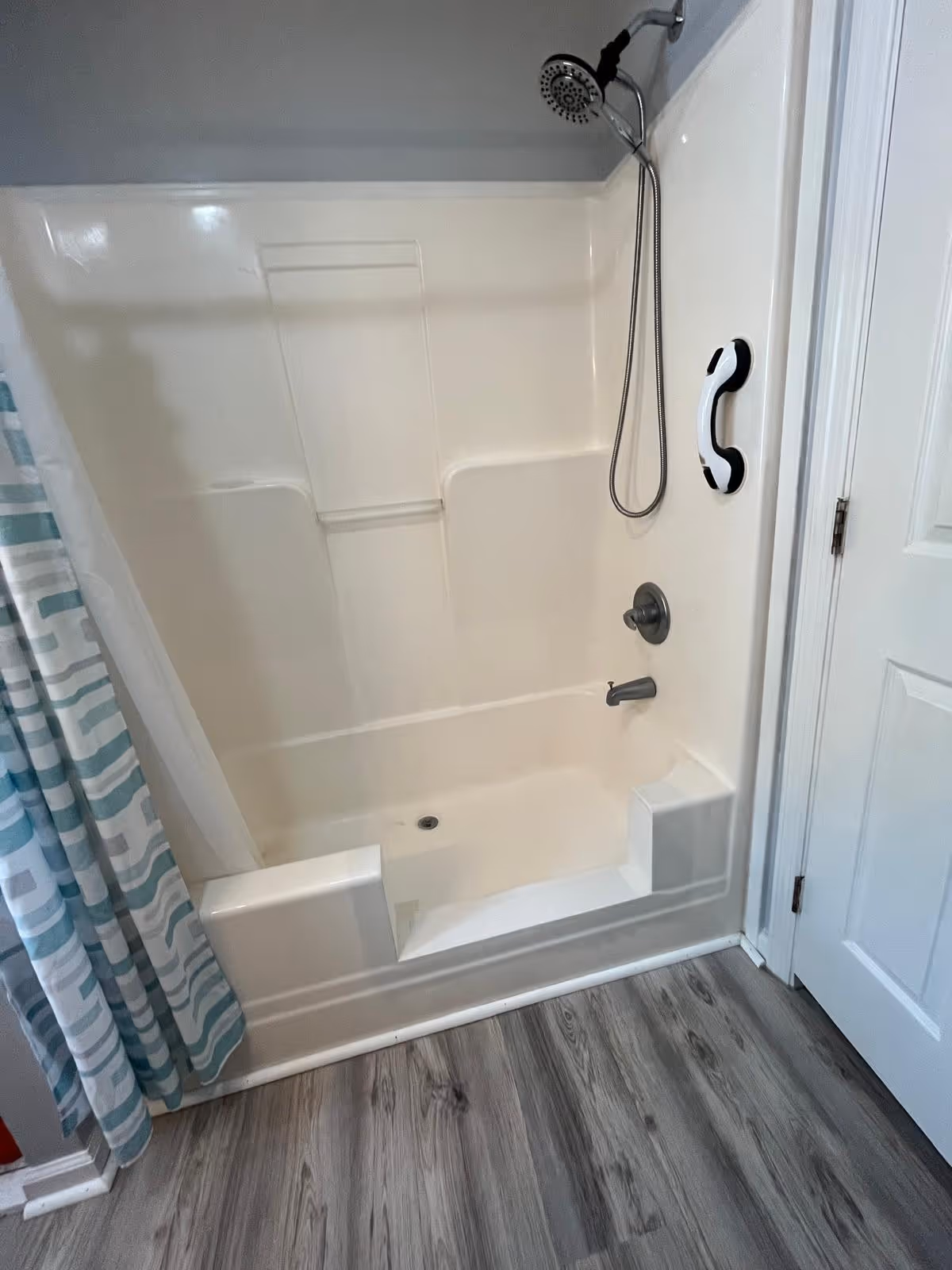 White bathtub with a showerhead, grab bar, and a striped shower curtain, next to a white door and gray wood floor.
