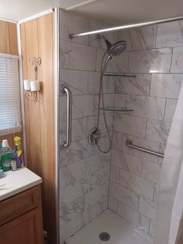 Shower corner with white marble-patterned tiles, a handheld showerhead, grab bars, and two glass corner shelves.