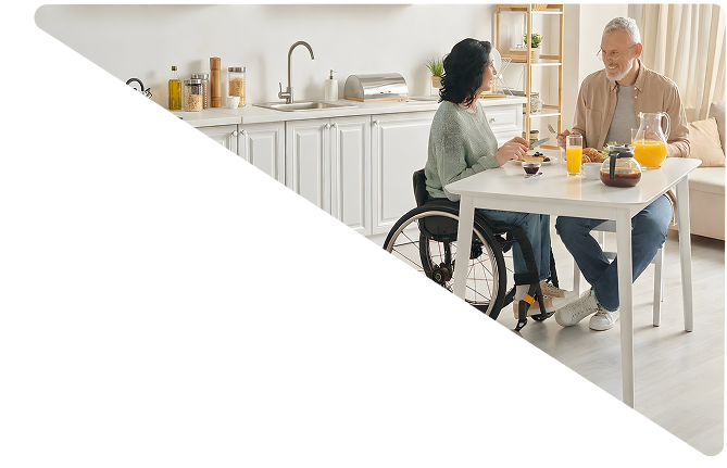 A man and woman in a wheelchair smiling and having breakfast together at a kitchen table with orange juice and coffee.