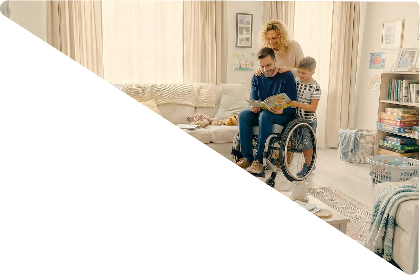 Family in a living room with a man in a wheelchair reading a book to a boy, and a woman standing behind them smiling.