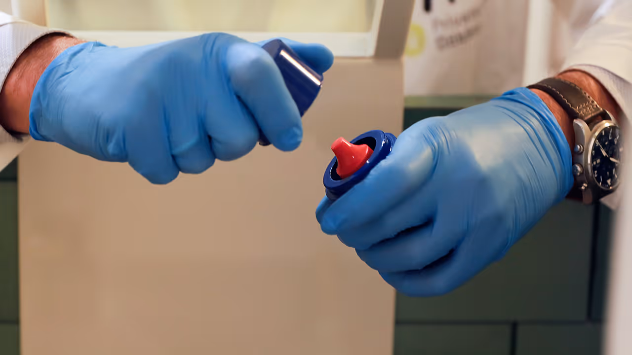 Dr. Markus Mitterhauser mit blauen Handschuhen öffnet einen blauen Kunststoffbehälter, in dem sich eine rotes Fläschchen befindet.