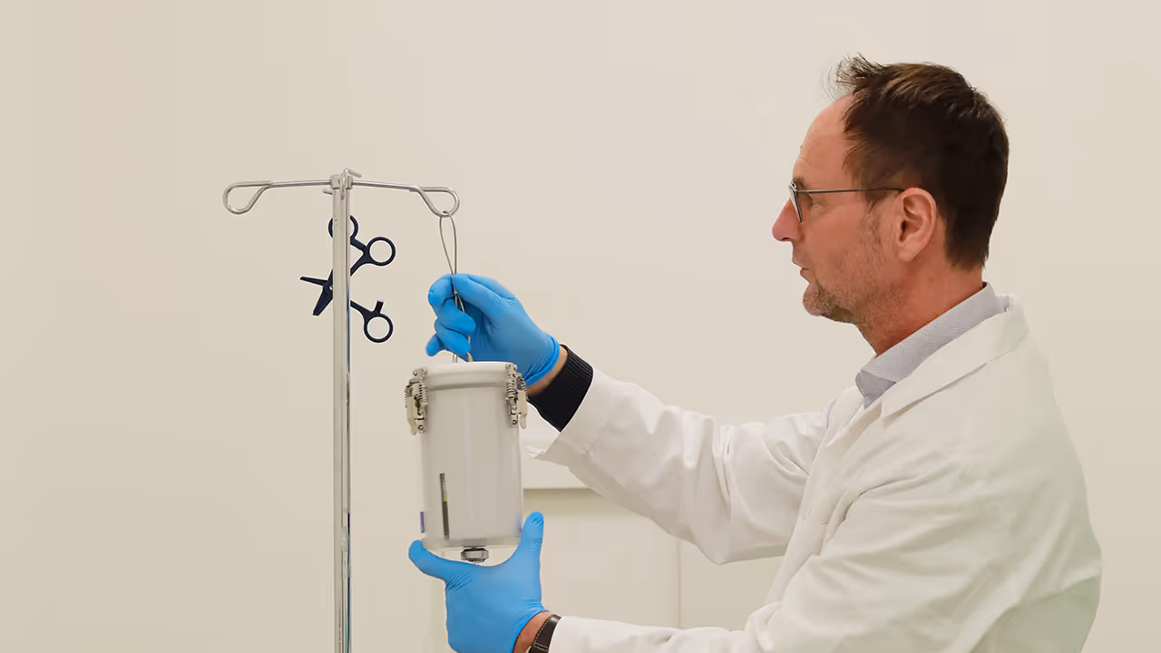 Dr. Markus Mitterhauser befestigt in einem Laborkittel und mit blauen Handschuhen einen weißen Behälter mit Metallinstrumenten an einem Infusionsständer in einem Untersuchungsraum.