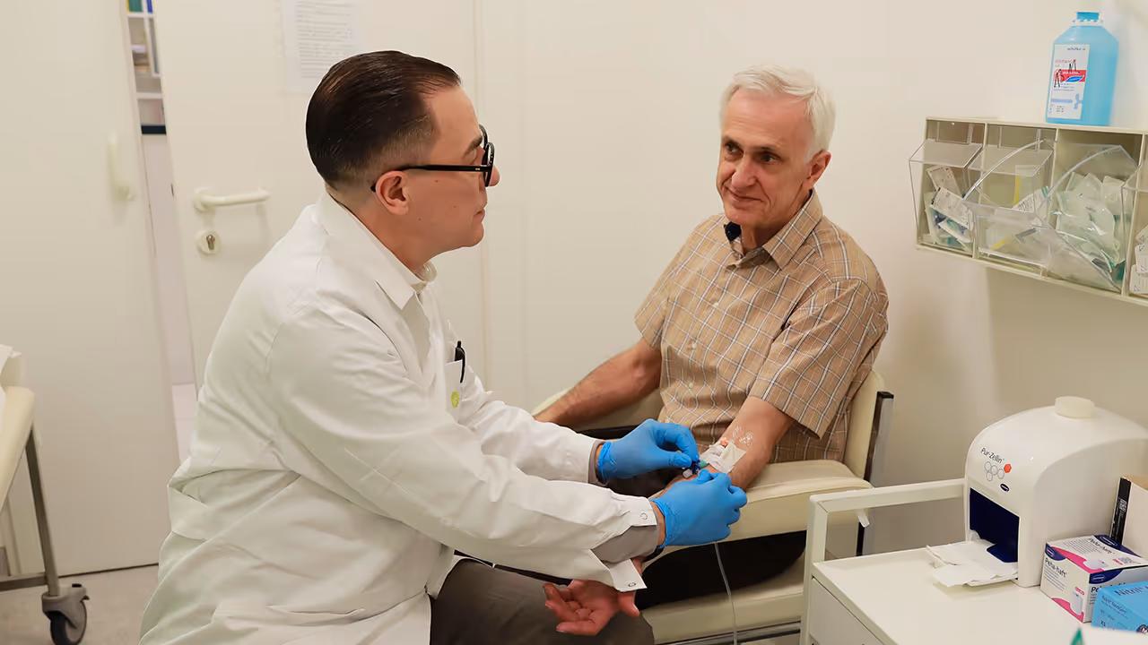Dr. Marcus Hacker, wearing a white coat and blue gloves, is seated with an older man in an examination room and is placing an intravenous line in his arm.