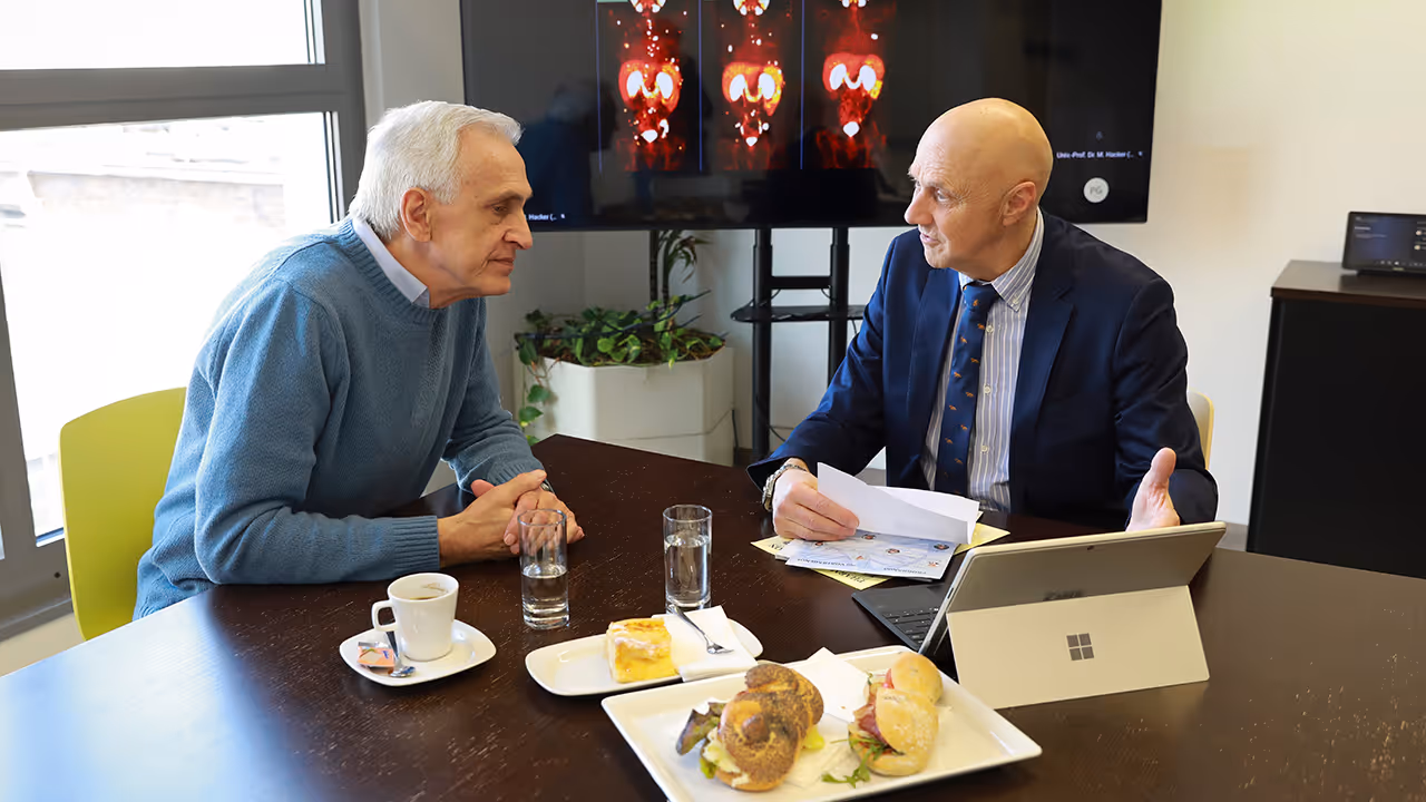 Dr. Werner Langsteger is seated with an older patient at a table with food, drinks, and documents, engaged in a conversation in a consultation room at Theranosticum Vienna; behind them, a medical image is displayed on a monitor.
