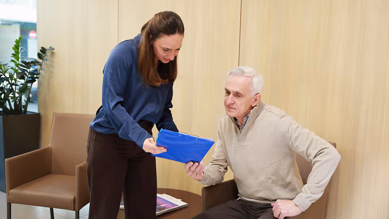 A staff member of Theranosticum Vienna shows documents in a blue folder to an older patient who is seated in a waiting area with chairs and a wooden wall.