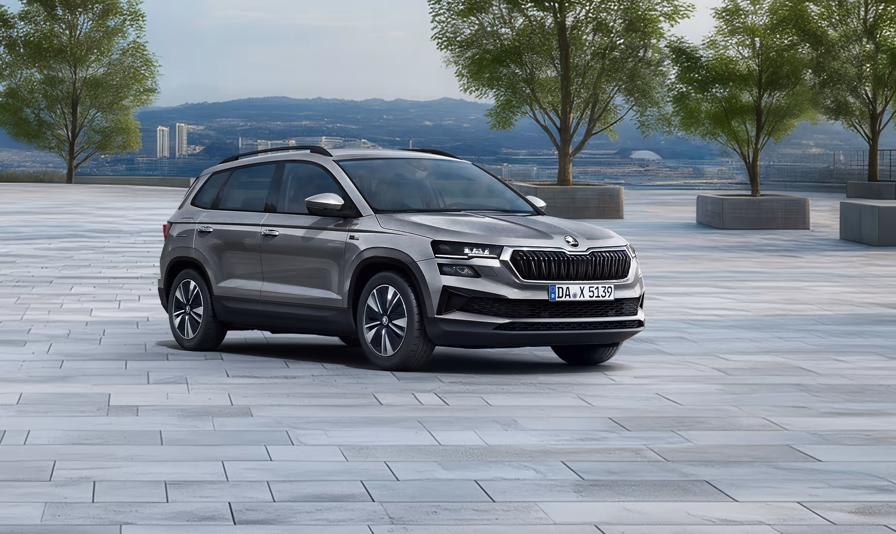 Front-Schrägansicht des Škoda Karoq Balance in der Lackierung Graphite-Grau Metallic auf einem gepflasterten Platz vor einer Stadtkulisse. Das SUV ist mit einer schwarzen Dachreling und dunklen, glanzgedrehten Leichtmetallfelgen ausgestattet. Im unteren Bildbereich weist ein grüner Kasten auf einen Preisvorteil von 2.900 € sowie die inkludierte 5-Jahres-Garantie hin.