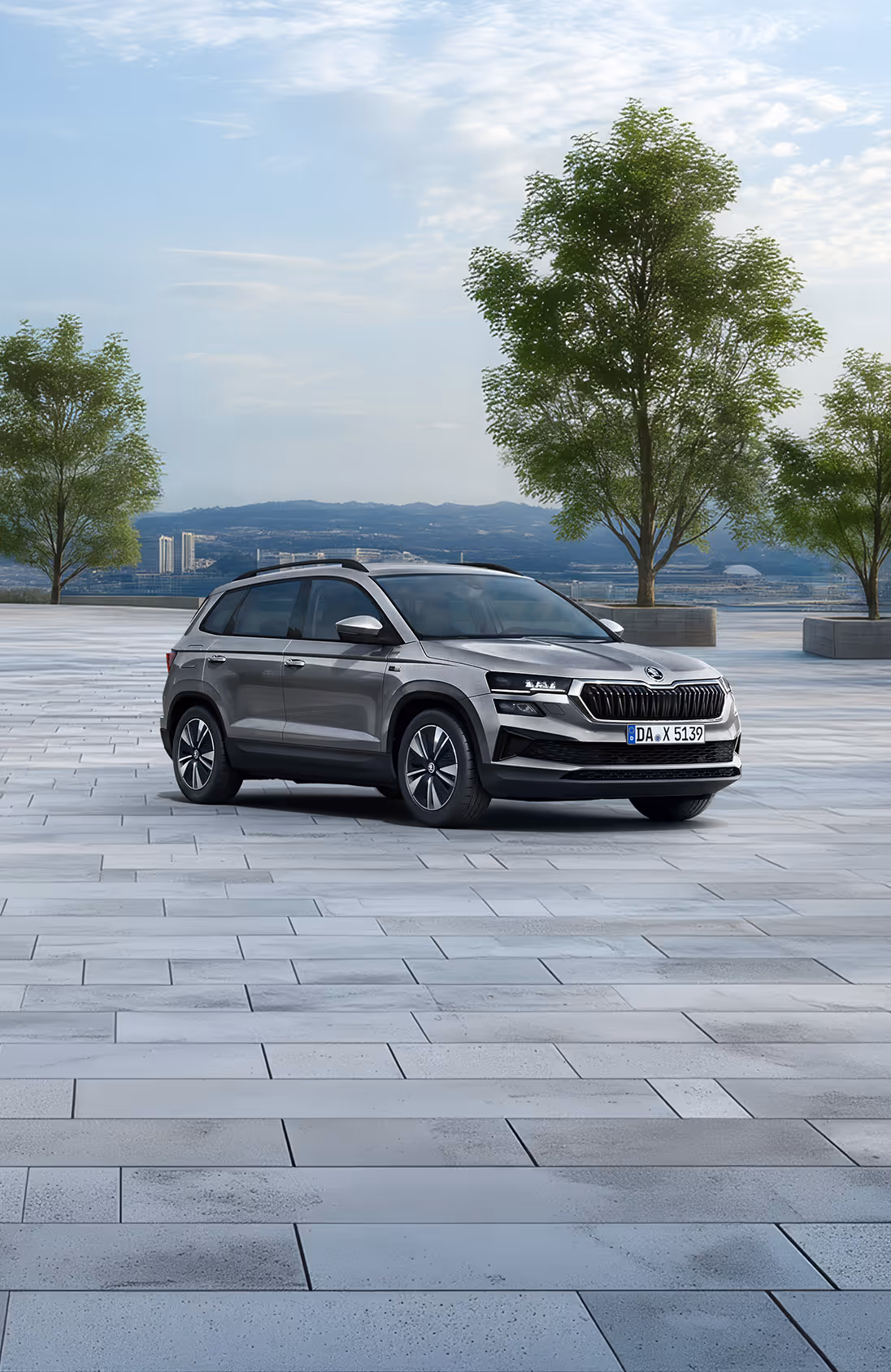 Front-Schrägansicht des Škoda Karoq Balance in der Lackierung Graphite-Grau Metallic auf einem gepflasterten Platz vor einer Stadtkulisse. Das SUV ist mit einer schwarzen Dachreling und dunklen, glanzgedrehten Leichtmetallfelgen ausgestattet. Im unteren Bildbereich weist ein grüner Kasten auf einen Preisvorteil von 2.900 € sowie die inkludierte 5-Jahres-Garantie hin.