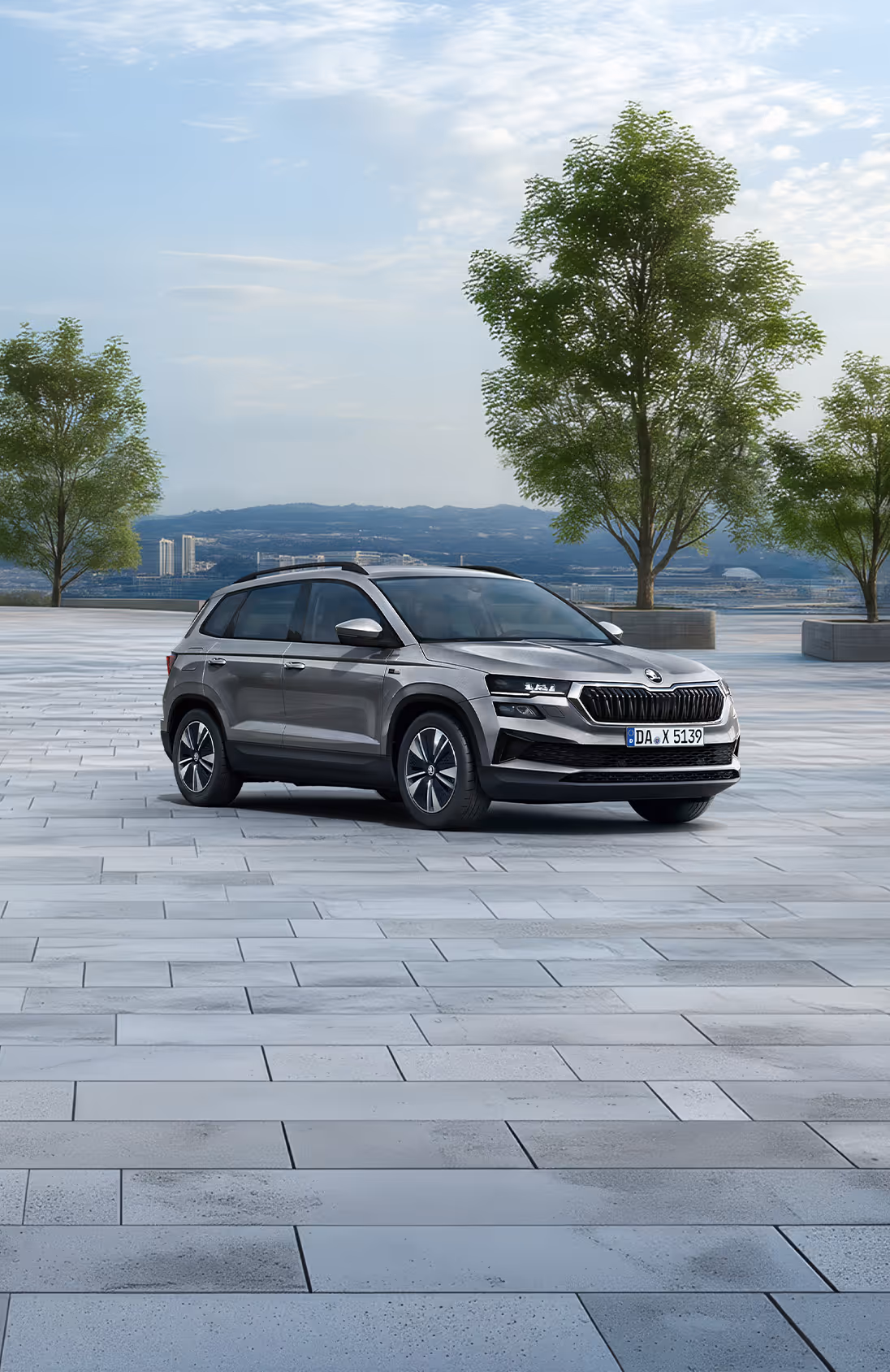 Front-Schrägansicht des Škoda Karoq Balance in der Lackierung Graphite-Grau Metallic auf einem gepflasterten Platz vor einer Stadtkulisse. Das SUV ist mit einer schwarzen Dachreling und dunklen, glanzgedrehten Leichtmetallfelgen ausgestattet. Im unteren Bildbereich weist ein grüner Kasten auf einen Preisvorteil von 2.900 € sowie die inkludierte 5-Jahres-Garantie hin.