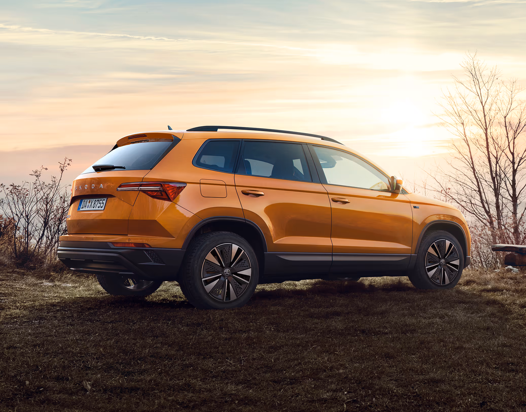 Heck-Schrägansicht des Škoda Karoq Balance in der Lackierung Phoenix-Orange Metallic in einer natürlichen Umgebung bei Sonnenuntergang. Das Bild zeigt das Fahrzeug auf unbefestigtem Untergrund, ausgestattet mit einer schwarzen Dachreling und dunklen Leichtmetallfelgen.