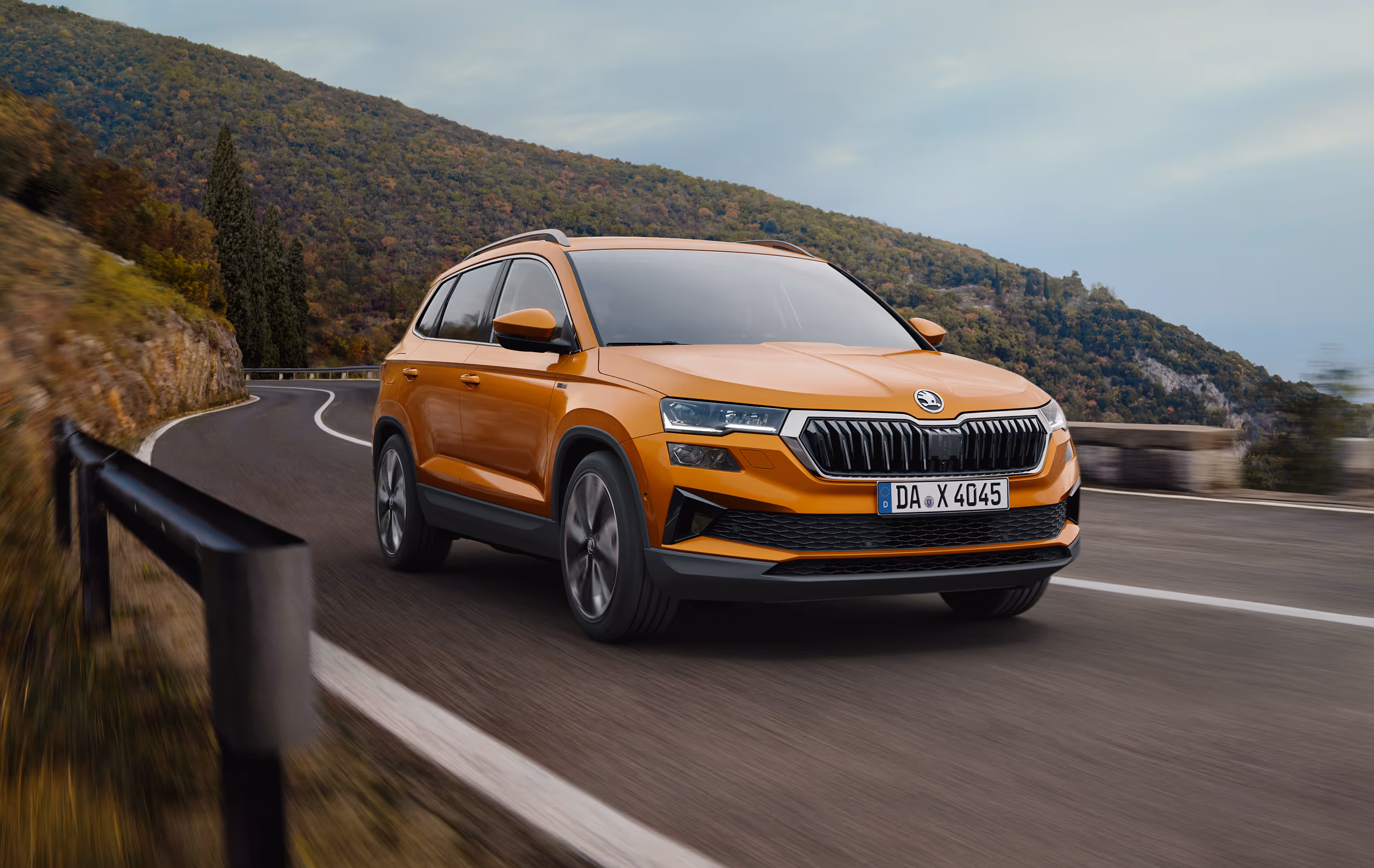 Ein Škoda Karoq Balance in der Lackierung Phoenix-Orange Metallic fährt auf einer kurvenreichen Straße entlang einer Felswand. Das Fahrzeug wird in einer dynamischen Dreiviertelansicht von vorne gezeigt, während die Bewegungsunschärfe des Hintergrunds die Fahrszene unterstreicht. Unten rechts steht auf einer dunkelgrün abgesetzten Fläche "Sicherheit & Assistenten" geschrieben.