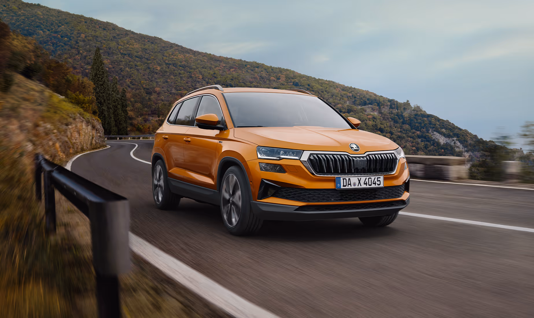 Ein Škoda Karoq Balance in der Lackierung Phoenix-Orange Metallic fährt auf einer kurvenreichen Straße entlang einer Felswand. Das Fahrzeug wird in einer dynamischen Dreiviertelansicht von vorne gezeigt, während die Bewegungsunschärfe des Hintergrunds die Fahrszene unterstreicht. 