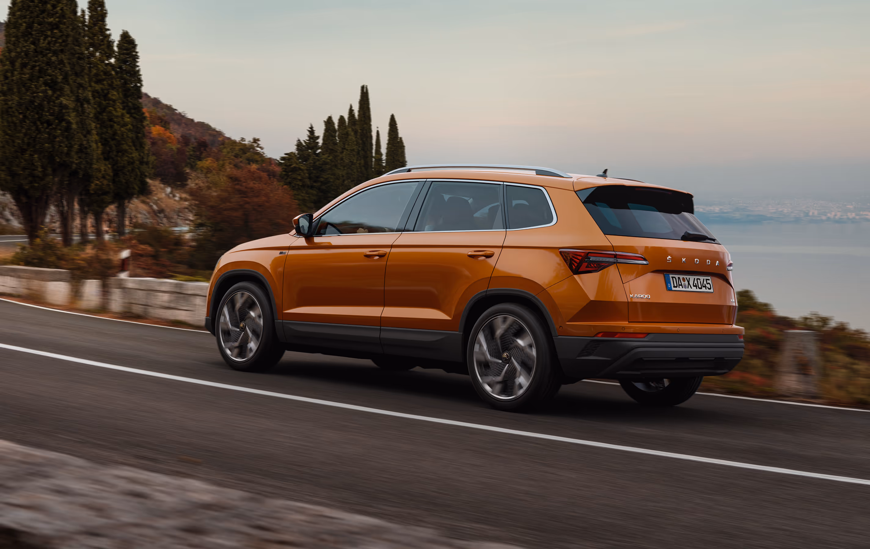 Ein Škoda Karoq Balance in der Lackierung Phoenix-Orange Metallic fährt auf einer kurvenreichen Küstenstraße. Das Fahrzeug wird von schräg hinten gezeigt, während es an Zypressen und einer hügeligen Landschaft mit Blick auf das Meer vorbeifährt.