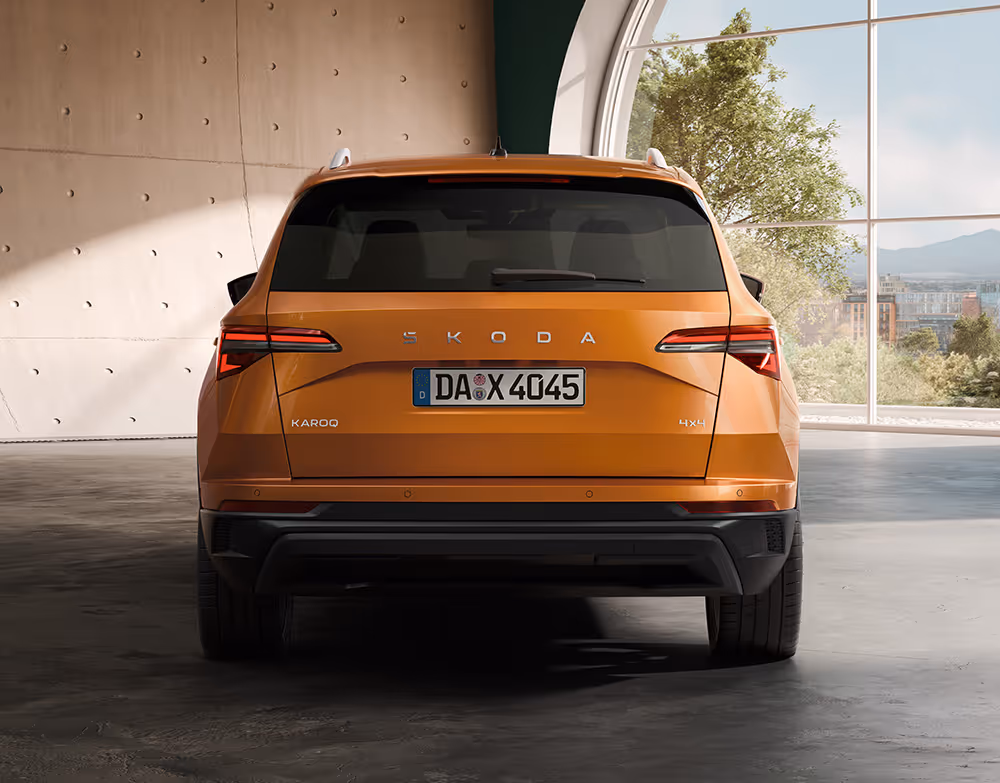 
Eine zentrale Heckansicht zeigt den Škoda Karoq Balance in der Lackierung Phoenix-Orange Metallic, platziert in einem modernen Raum mit Betonwänden und großem Rundbogenfenster. Im Fokus stehen der Škoda-Schriftzug auf der Heckklappe, die markanten Rückleuchten sowie die Modell- und 4x4-Schriftzüge. Das einfallende Licht hebt zudem die silberne Dachreling und den robusten schwarzen Heckstoßfänger hervor.