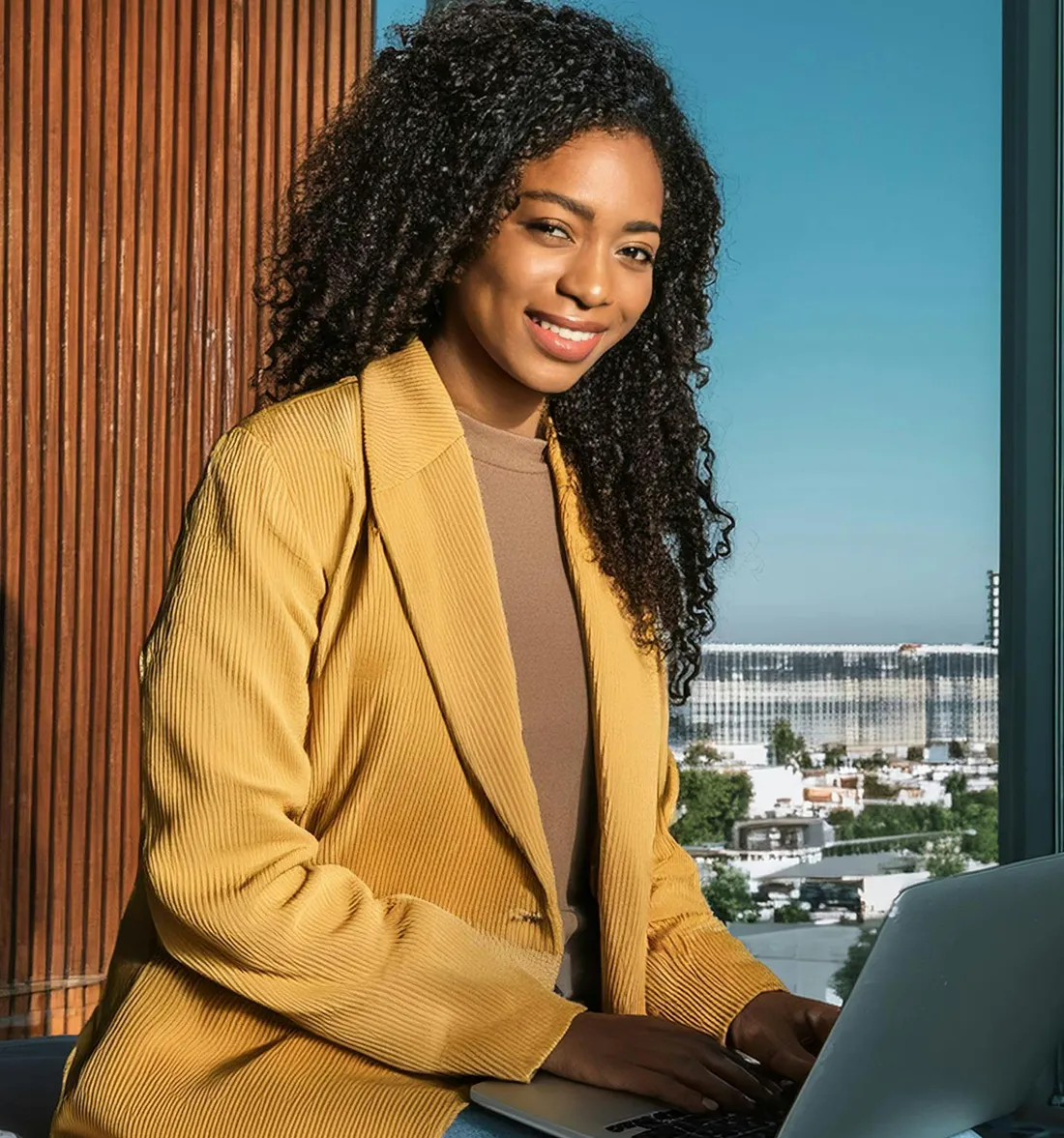 Smiling woman with curly hair wearing a yellow blazer working on a laptop by a window with a city view.