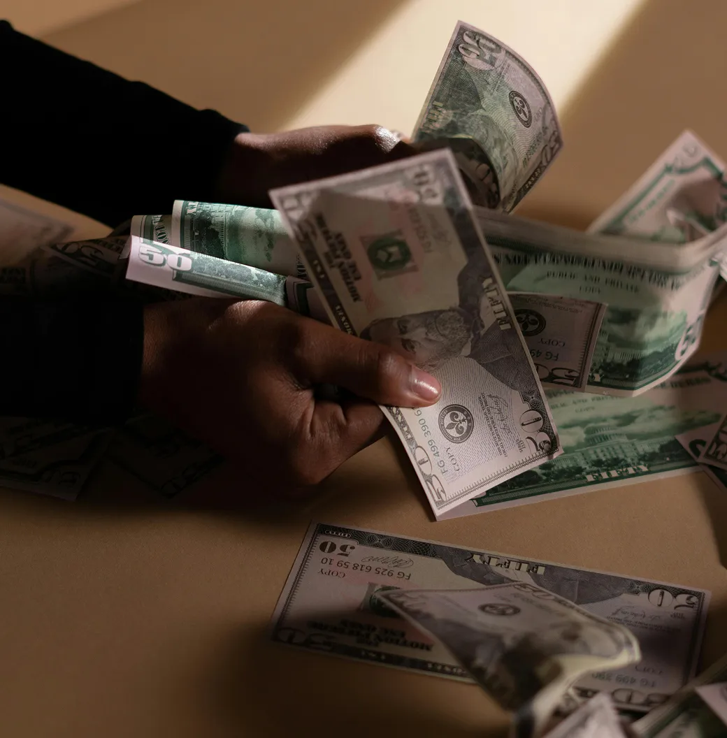 Hands holding and counting multiple US fifty-dollar bills over a beige surface.