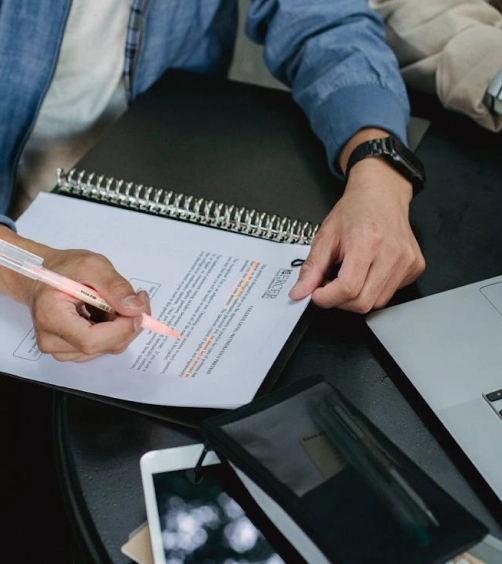 Person highlighting text on a printed document with a pink highlighter at a black table next to a laptop and smartphone.