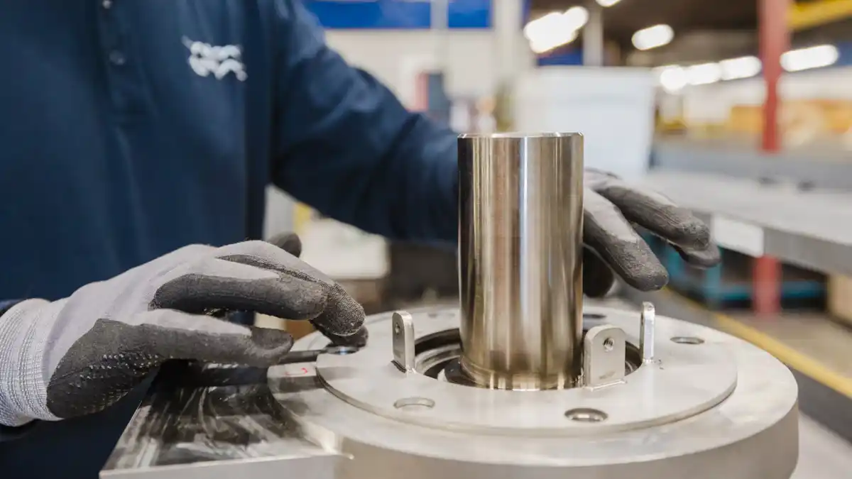 En arbeidstaker med hansker håndterer en metallkomponent i en industrifabrikk.