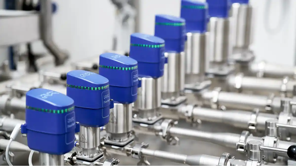 Rekke av blå automatiserte ventiler montert på rustfrie stålrør i et industrielt anlegg.