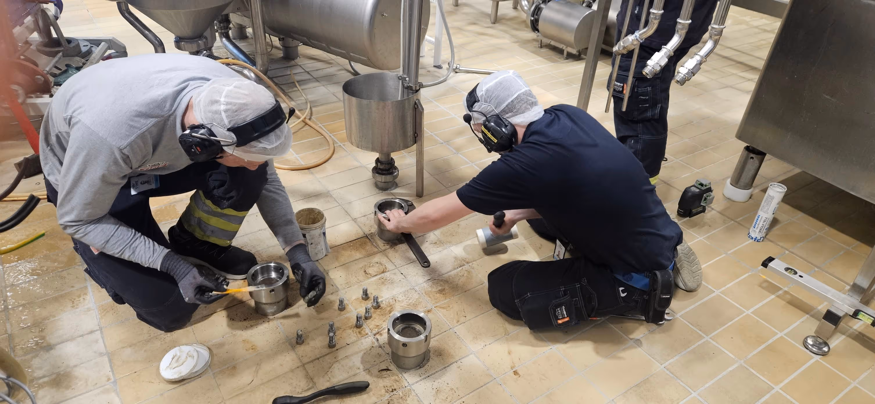To arbeidere med vernehodeutstyr og hørselvern utfører vedlikehold på stålmaskin deler på fabrikkgulv.