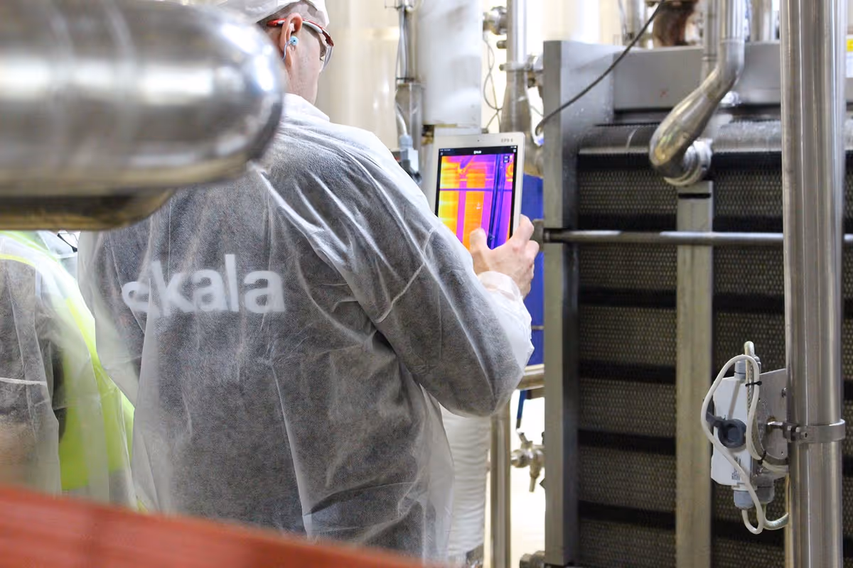 Person i vernedrakt holder et termisk kamera og utfører en inspeksjon i et industrilokale med rør og maskiner.