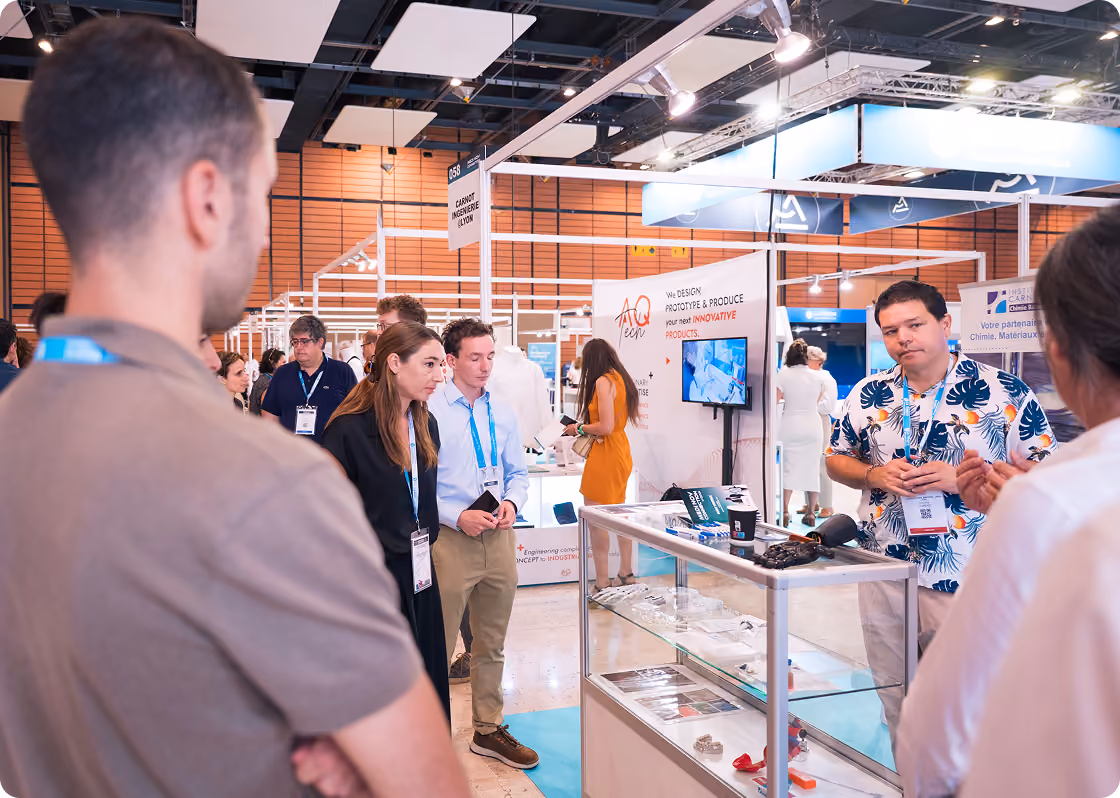 Des visiteurs discutent avec un exposant à un stand lors d'un salon professionnel, avec des badges d'identification visibles, des vitrines d'objets exposés et un écran affichant des informations.