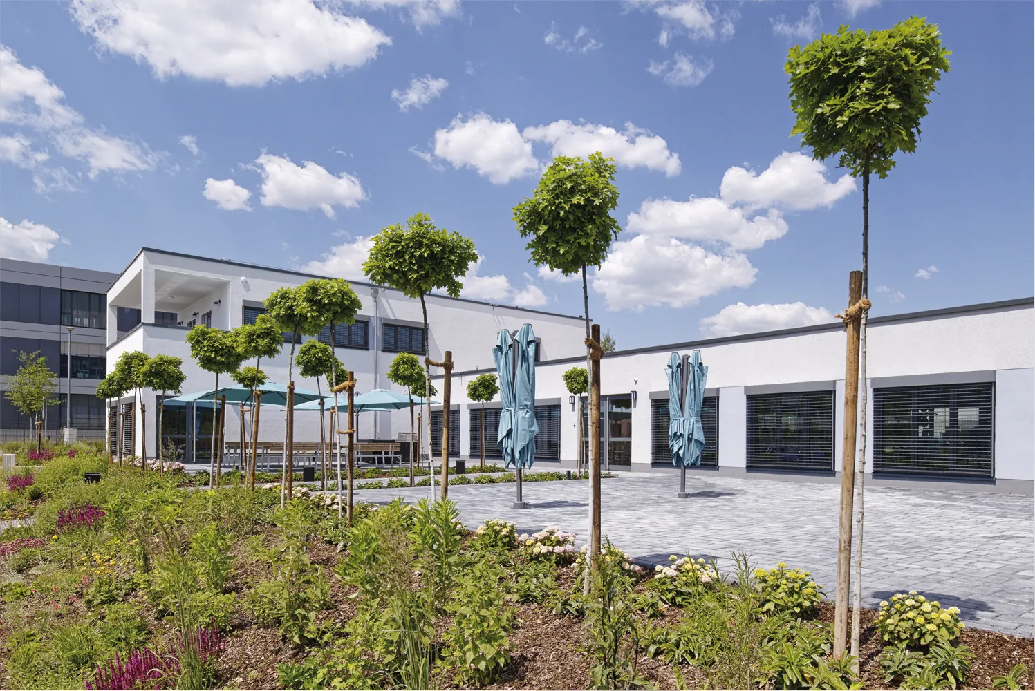 Modernes Bürogebäude mit jungen Bäumen, Terrasse mit blauen Sonnenschirmen und klar blauem Himmel.