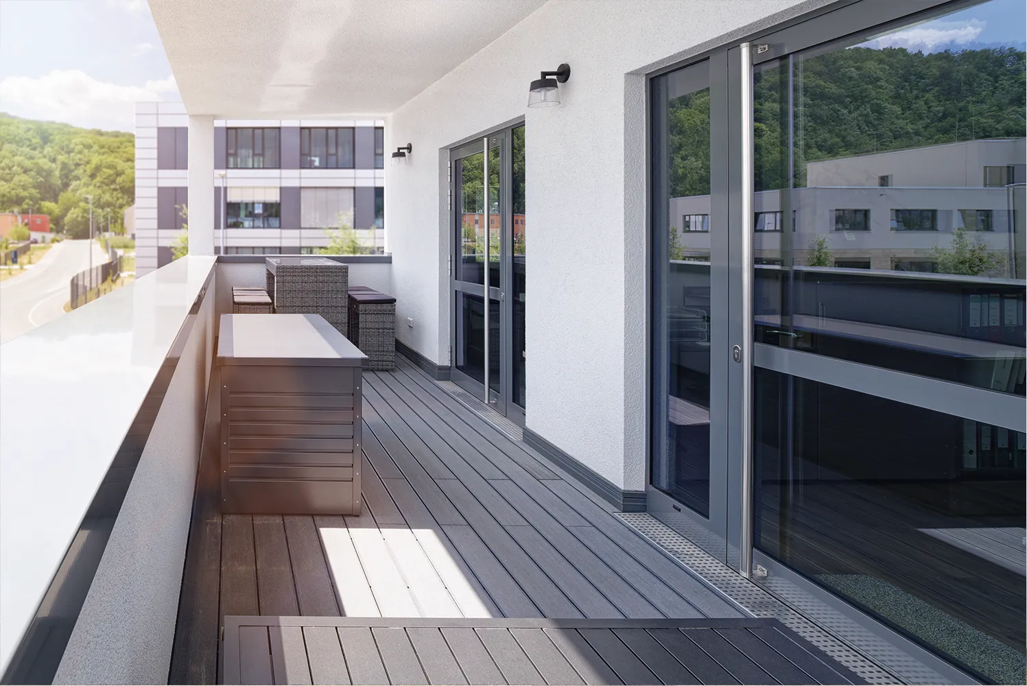 Moderner Balkon mit grauem Bodenbelag, Sitzgruppe aus Korbgeflecht und großen Glastüren mit Blick auf grüne Hügel und ein Bürogebäude.