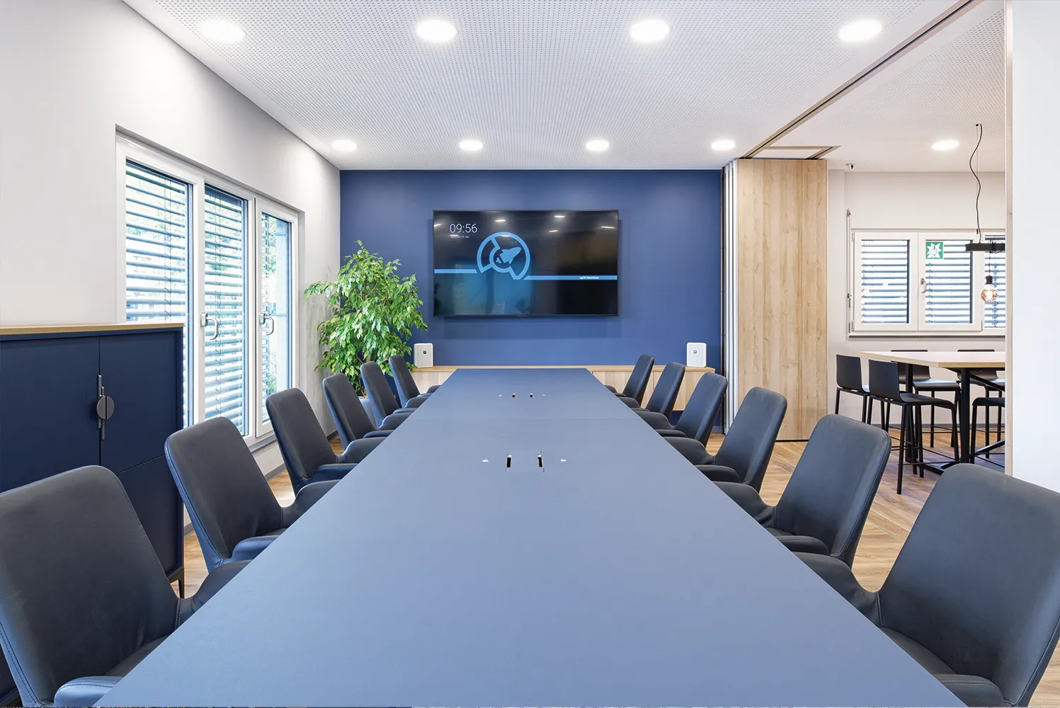 Moderner Besprechungsraum mit langem blauem Tisch, schwarzen Stühlen, großem Bildschirm und Pflanzen.