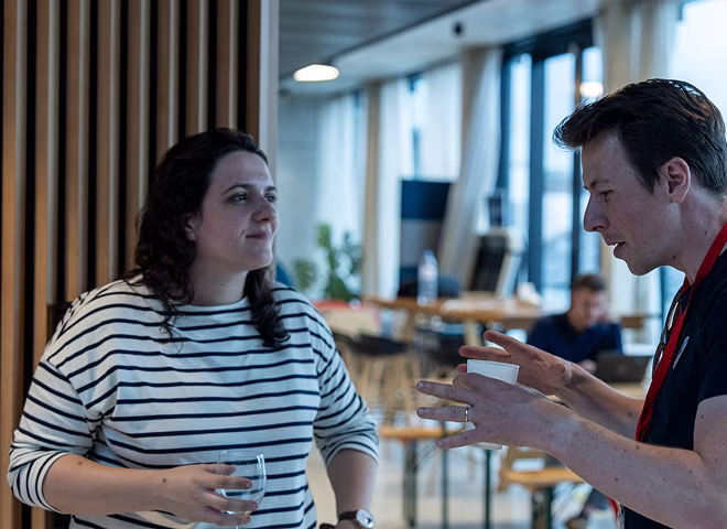 Deux personnes discutant dans un espace de bureau moderne, l'une tient un verre et l'autre une tasse en plastique.