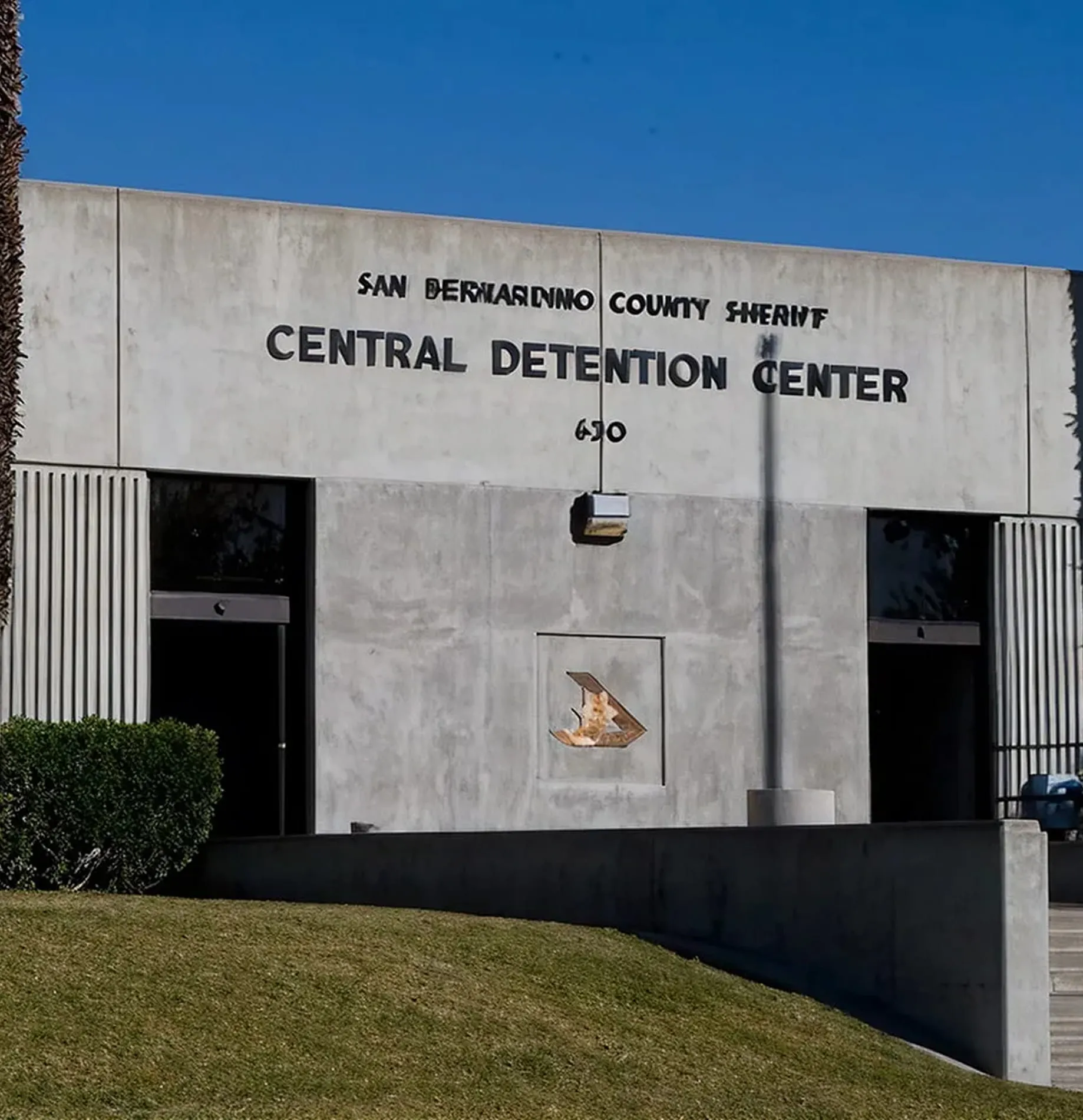 Exterior of the San Bernardino County Sheriff Central Detention Center with concrete walls, two entrance doors, and a patch of grass in front.