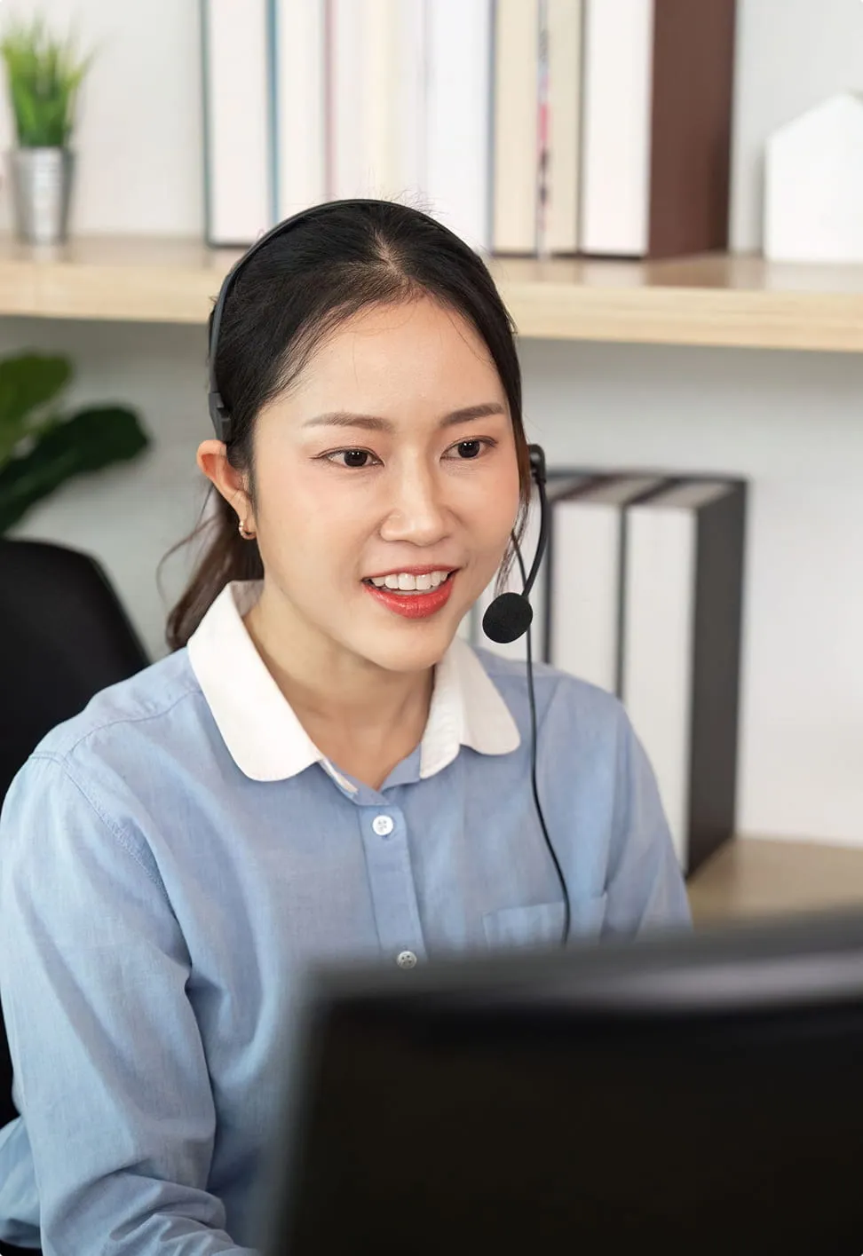 Woman wearing headset and blue shirt smiling while looking at a computer screen.