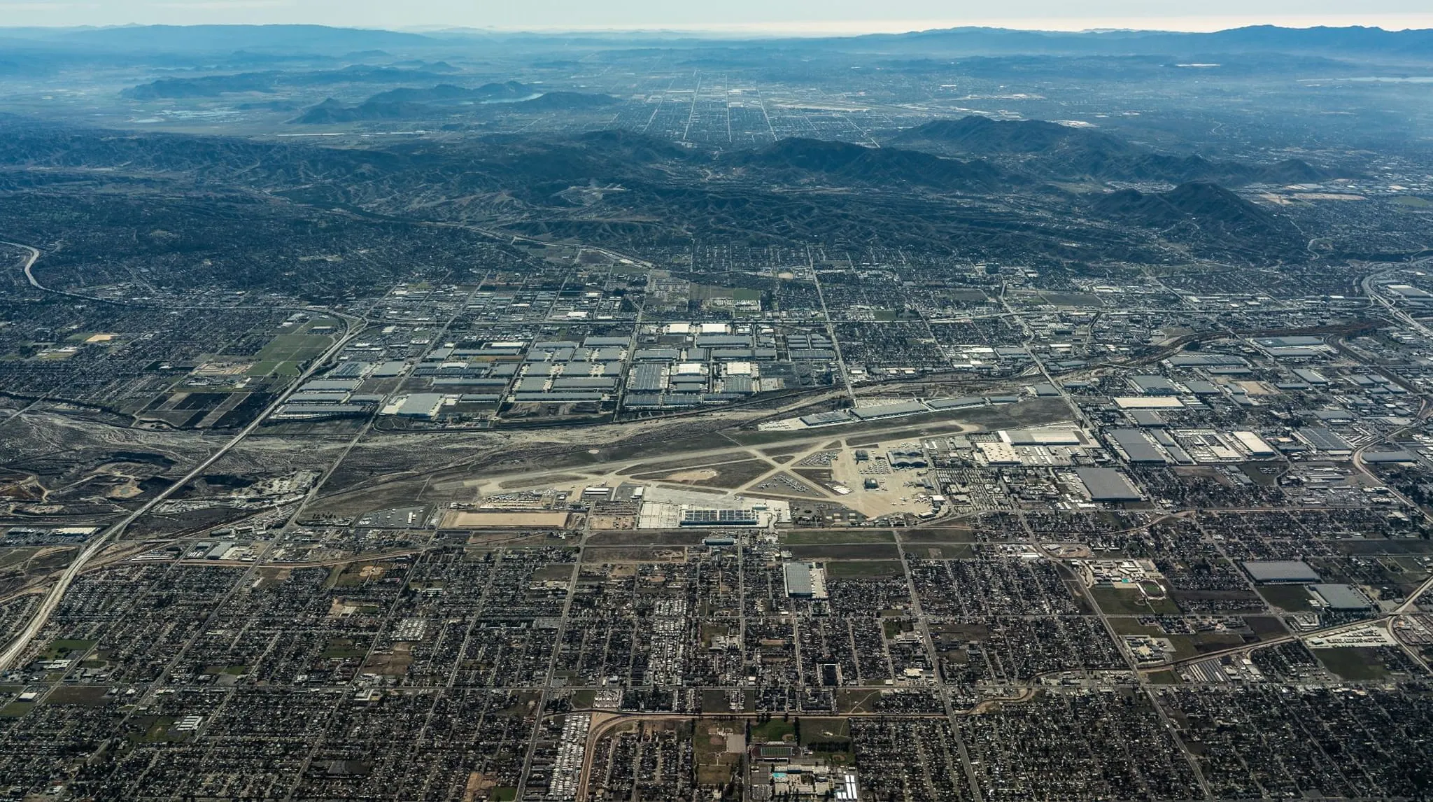 Aerial view of an urban area with an airport runway surrounded by industrial buildings and residential neighborhoods, with mountains in the background.