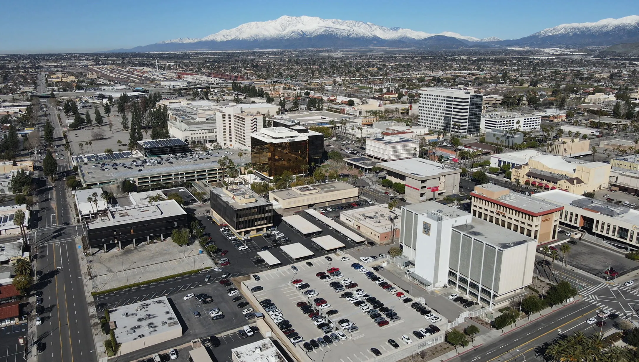 Aerial view of a cityscape with multiple buildings, parking lots filled with cars, and snow-covered mountains in the background under a clear blue sky.
