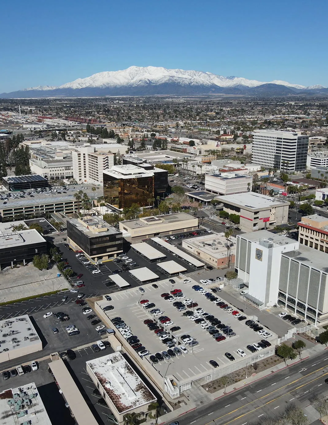 Aerial view of a city with a parking lot, mid-rise buildings, and snow-capped mountains under a clear blue sky.