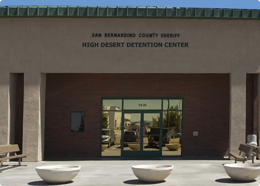 Entrance of San Bernardino County Sheriff High Desert Detention Center with glass doors reflecting cars and buildings.