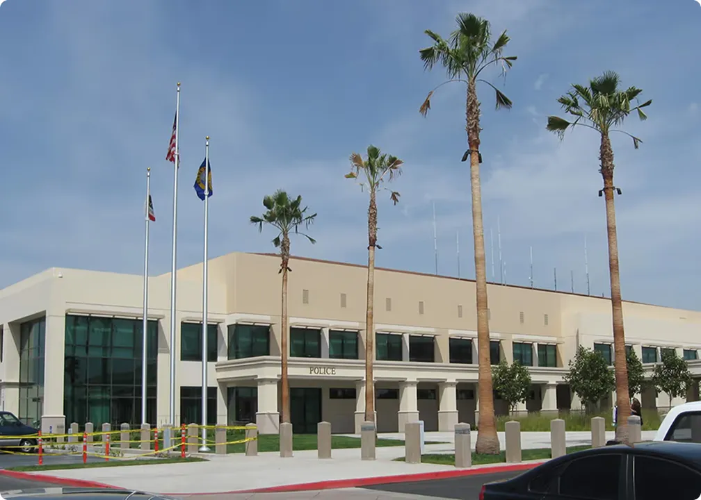 Modern police station building with palm trees, flagpoles displaying American and state flags, and a clear blue sky.