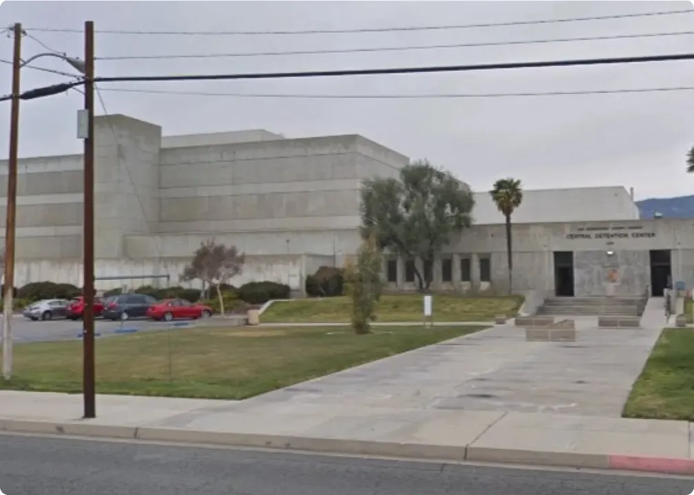 Exterior of a large concrete building labeled Central Detention Center with a grassy lawn, trees, and parked cars in front.