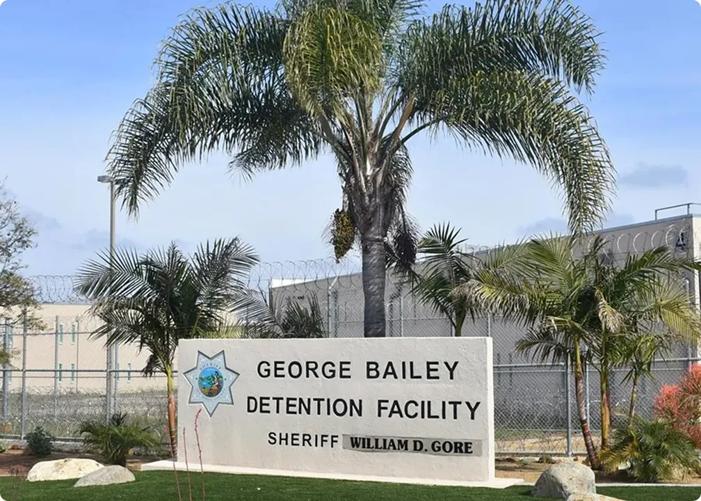 Sign for George Bailey Detention Facility with sheriff William D. Gore's name, palm trees, and barbed wire fencing in the background.