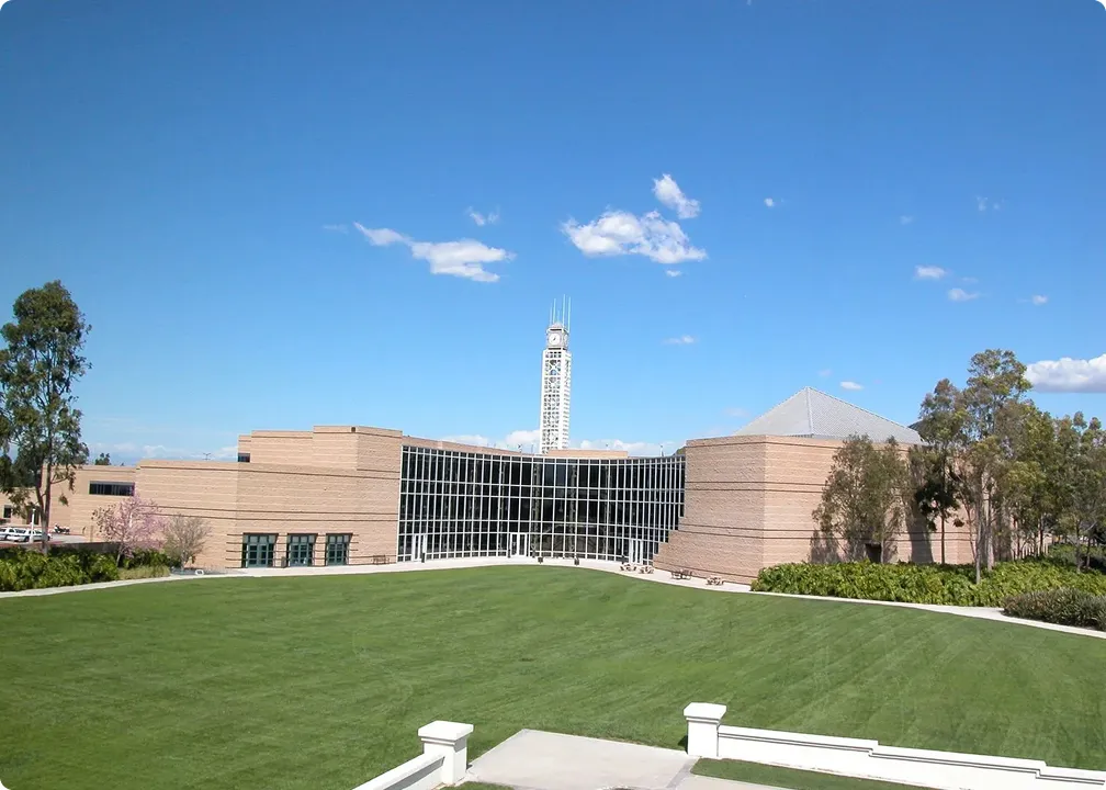 Modern beige brick building with large glass windows, a tall clock tower, and a green lawn in the foreground under a blue sky.